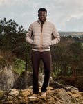 Man wearing a beige puffer jacket and dark pants standing on rocky terrain with trees and sky in the background
