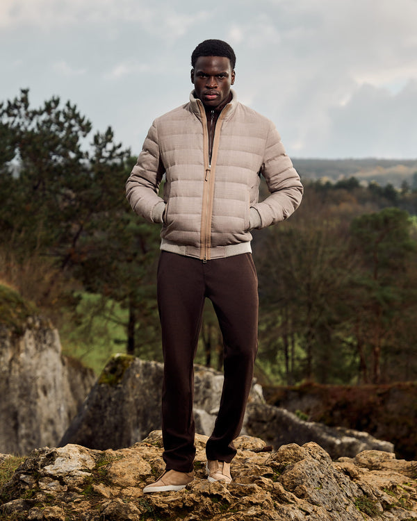Man wearing a beige puffer jacket and dark pants standing on rocky terrain with trees and sky in the background