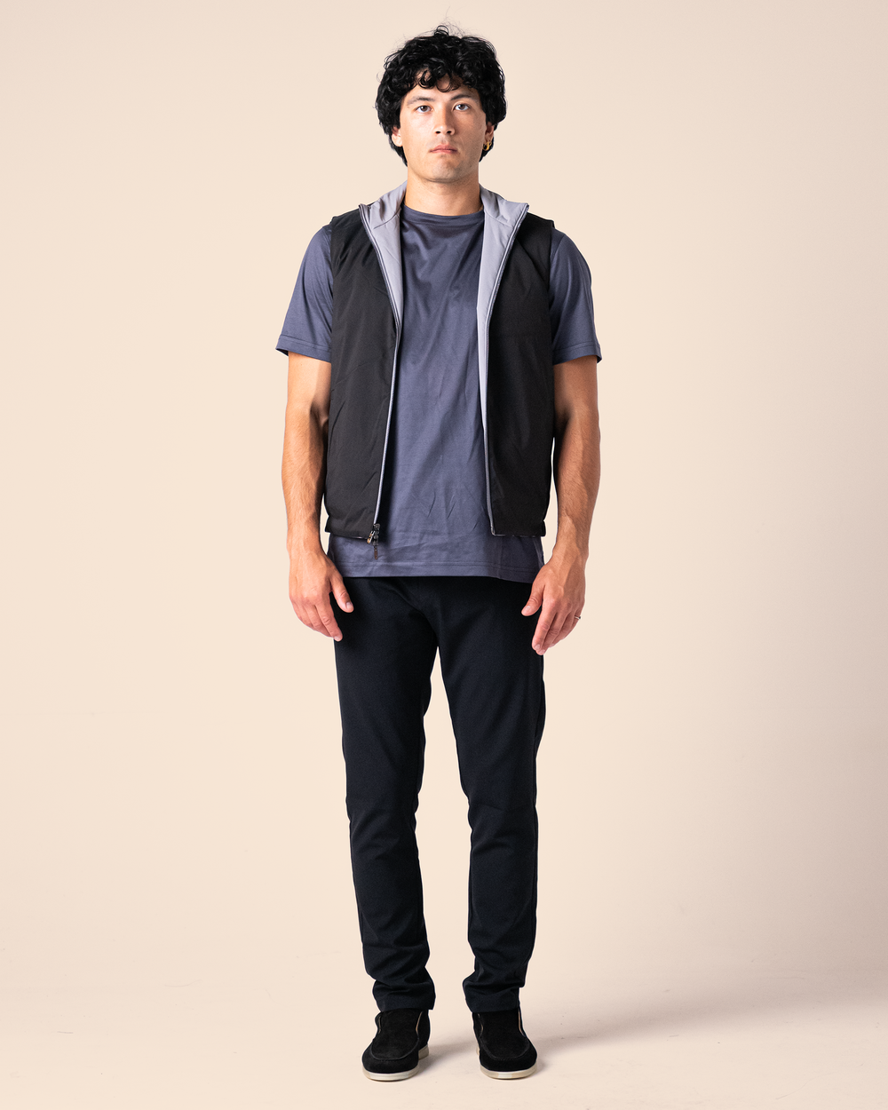 Man wearing a dark vest over a gray shirt with black pants on a beige background